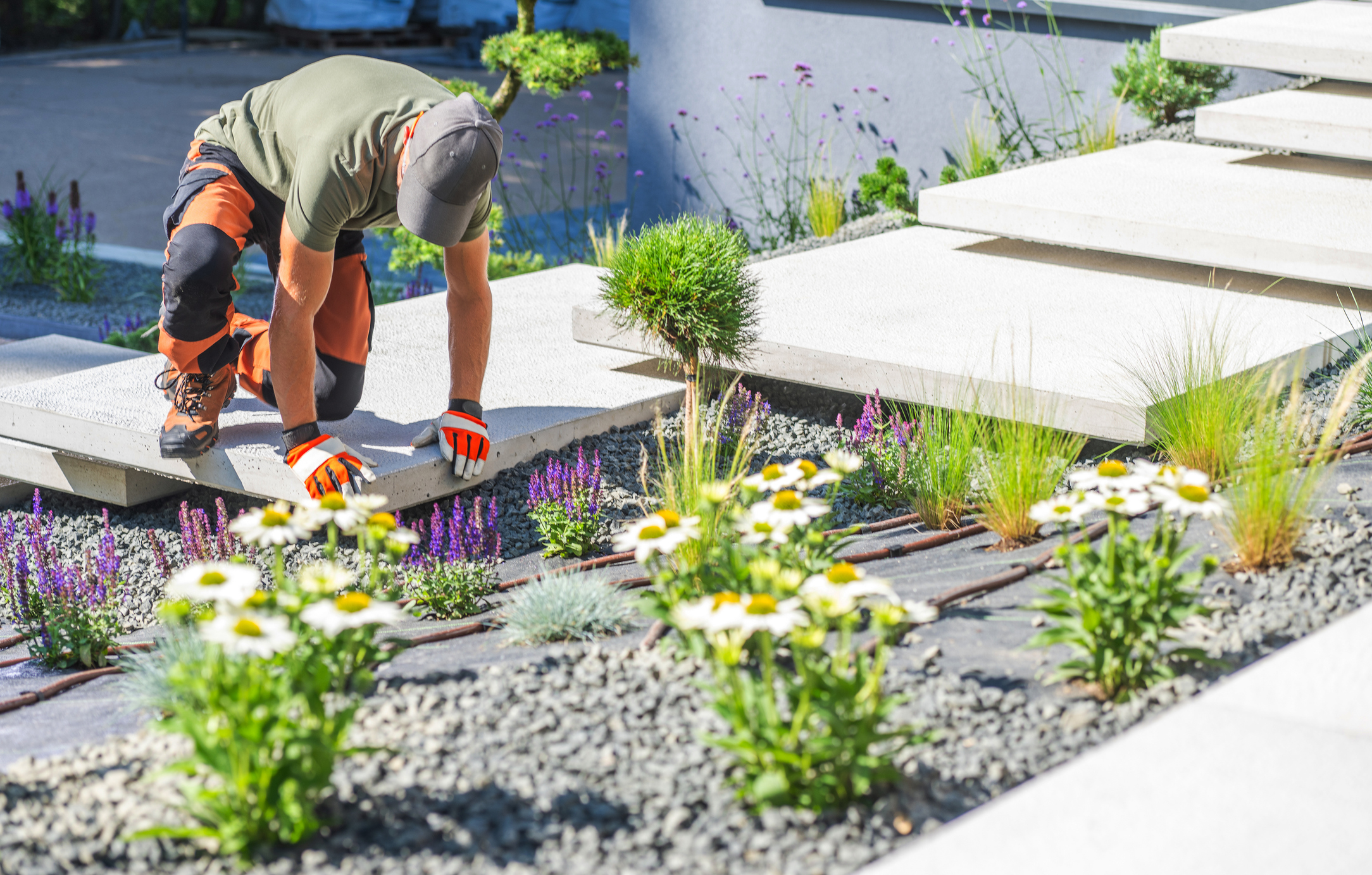 A gardener tends to vibrant flowers in a contemporary garden, kneeling on a stone pathway surrounded by colorful blooms under clear blue skies.